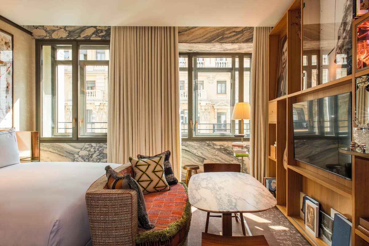 A hotel room with a large bed, a sofa with patterned cushions, a marble-topped table, a television, and a view of buildings through large windows.