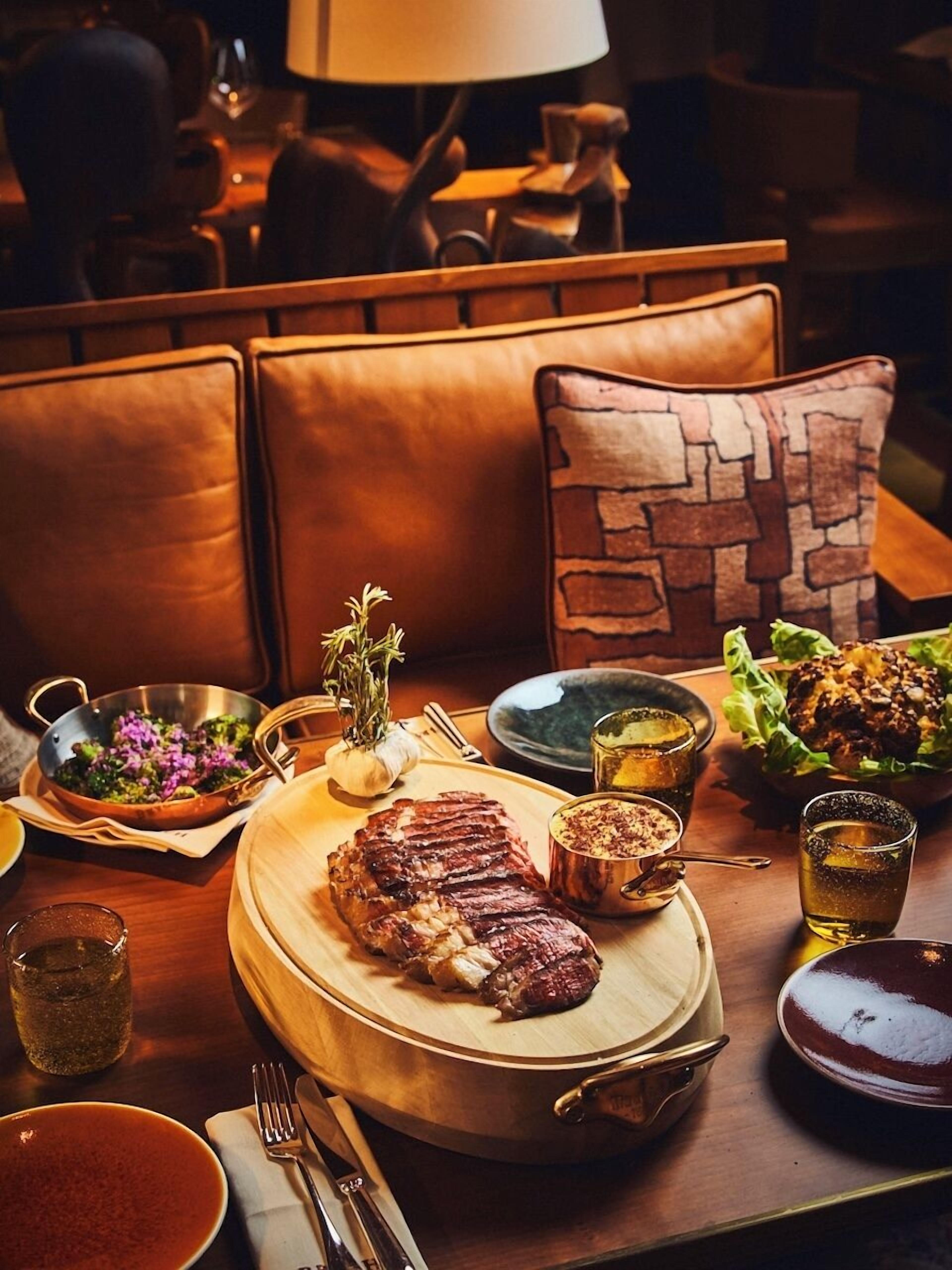 A dining table set with a steak, vegetables, and drinks, surrounded by leather chairs and a stone-patterned cushion.