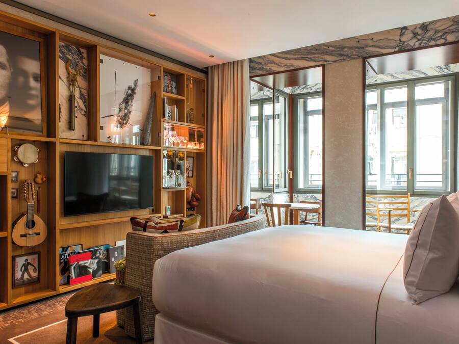 A hotel room with a large bed, a television, a wooden bookshelf, and a view of the city through large windows.