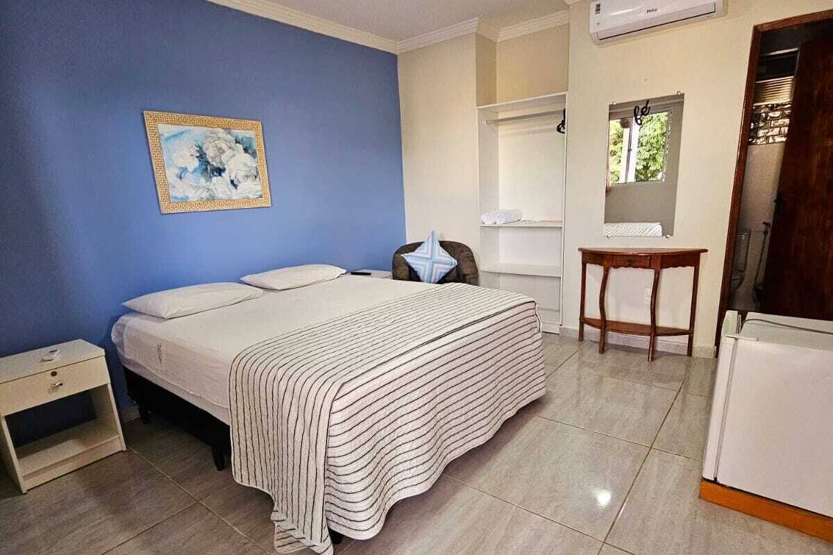 Un dormitorio con cama, mesita de noche, silla, una pequeña mesa y un aire acondicionado.