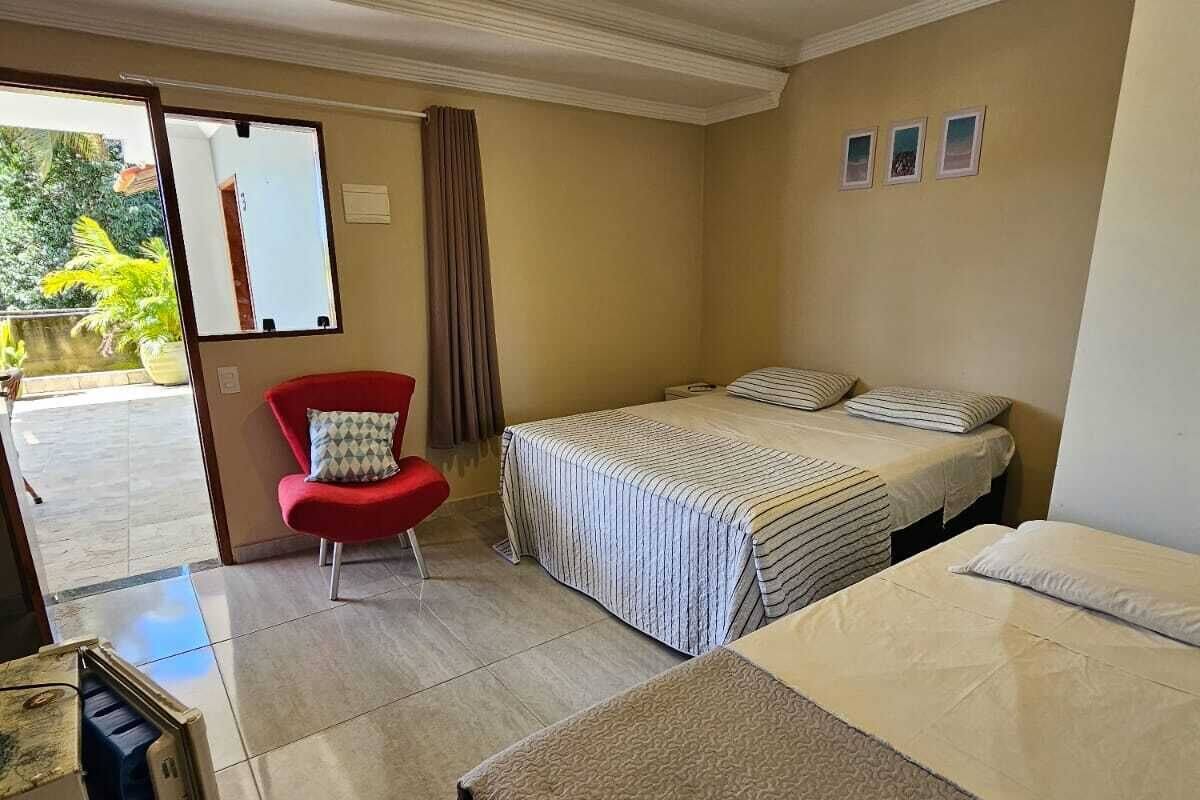 Habitación de hotel con dos camas, una silla roja, una mesita y vistas al exterior con vegetación.