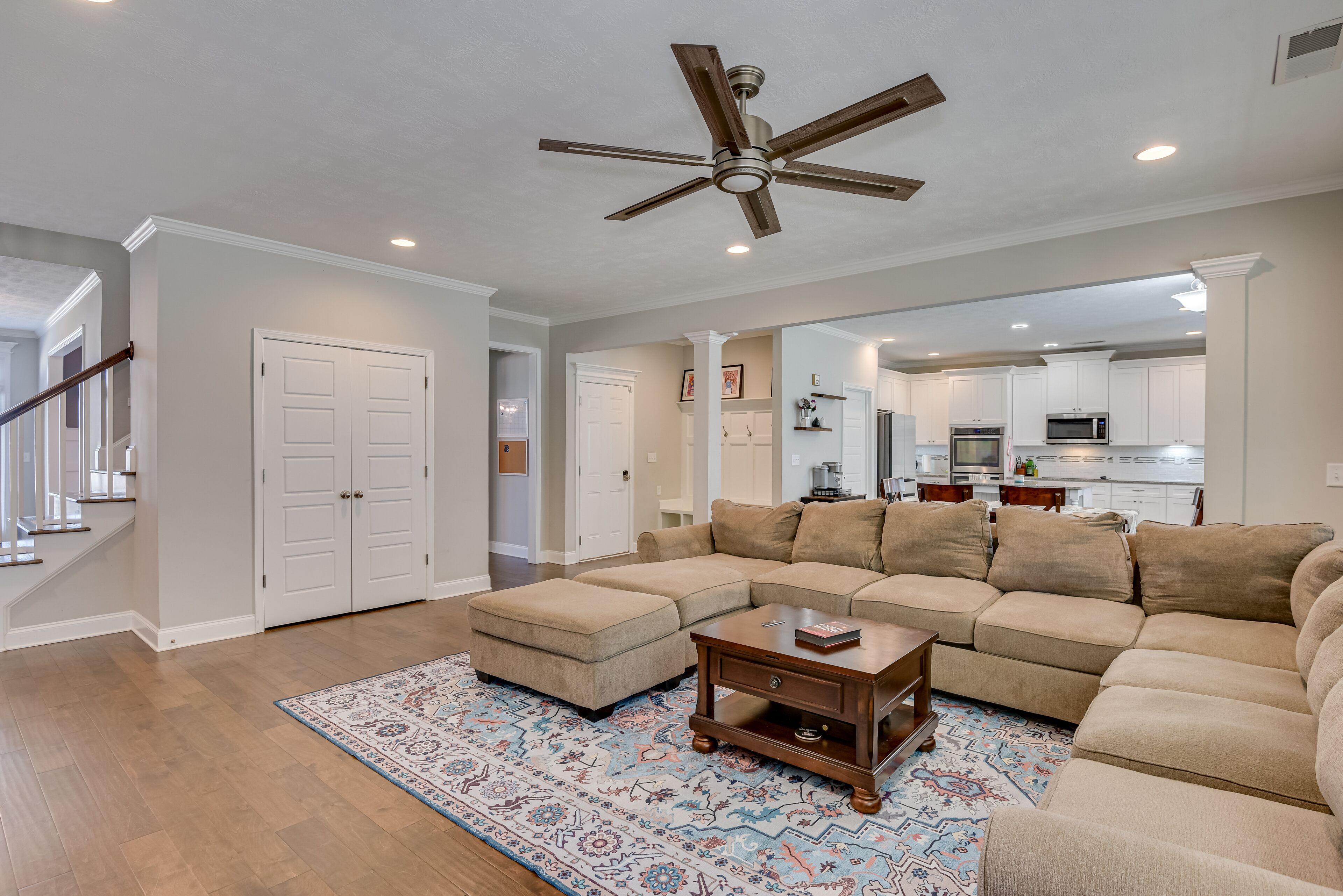 A spacious living room with a sectional sofa, ottoman, and a coffee table. A ceiling fan and recessed lighting are visible.