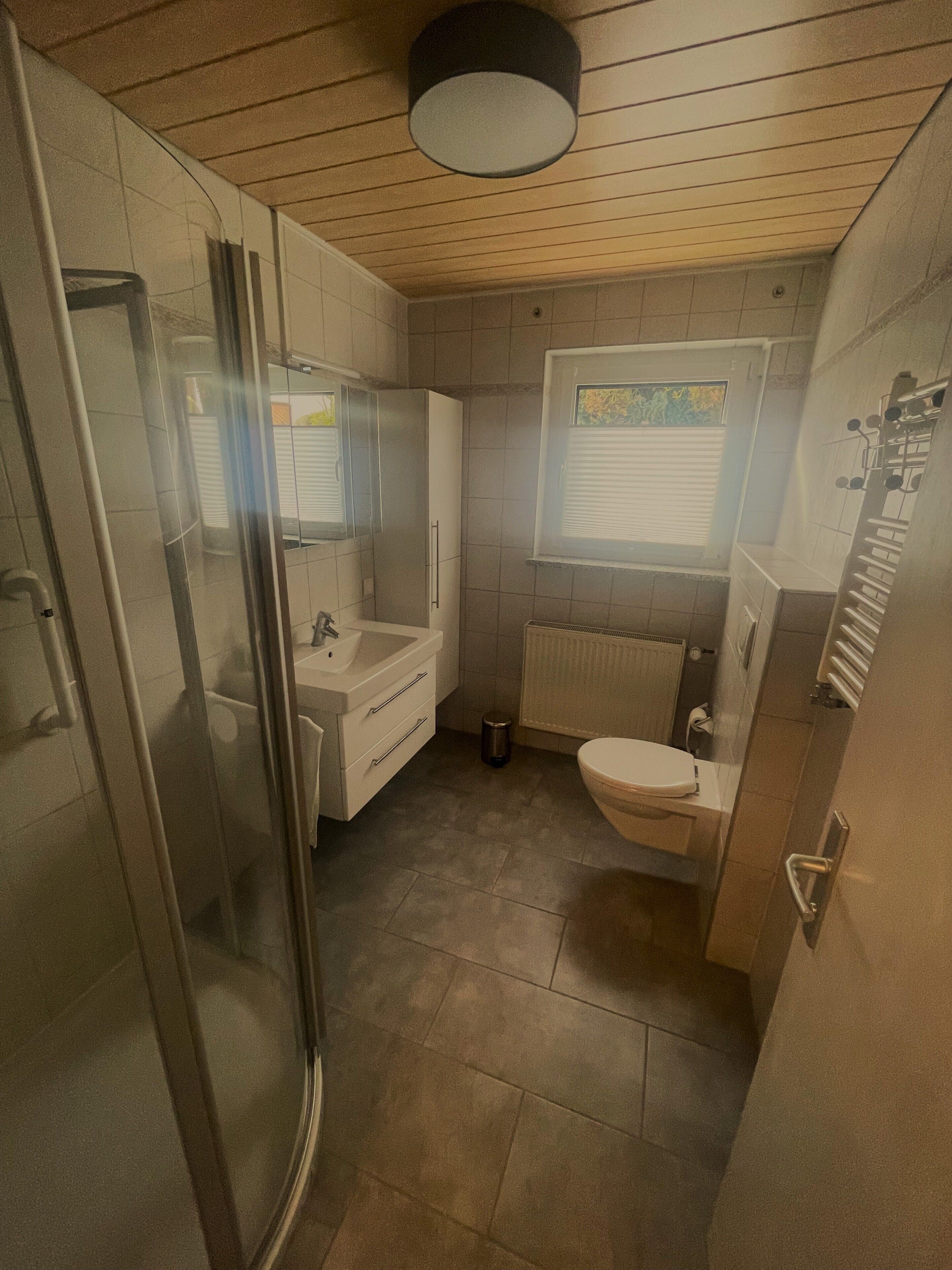A compact bathroom with a glass shower enclosure, a sink, a toilet, and a window with blinds.