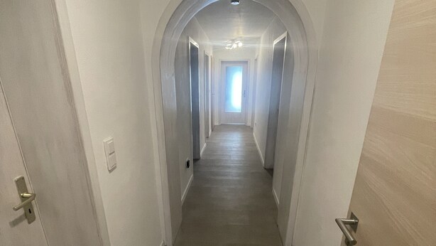 En smal, väl upplyst hall med trägolv och vita väggar, som leder fram till en dörr i slutet.