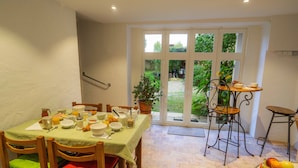 A dining area with a table set for breakfast, chairs with cushions, a potted plant, and a view through a glass door to the outside.