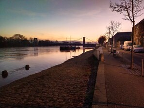 A riverside walkway with a cobblestone path, a bridge in the background, and parked cars along the side. - Les Balcons sur la Loire (Chalonnes-sur-Loire)