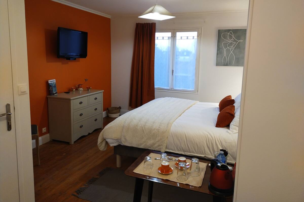 Une chambre à coucher comprenant un lit, une commode, un téléviseur, une fenêtre avec des rideaux et une table sur laquelle se trouvent une bouilloire et des tasses.