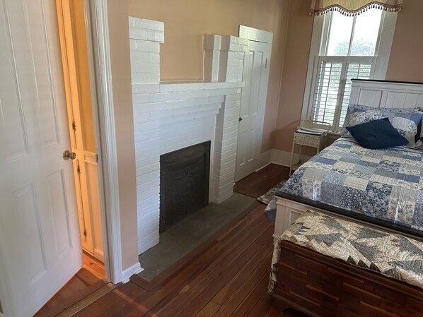 A bedroom with a white brick fireplace, wooden flooring, a bed with a quilt, and a window with curtains.