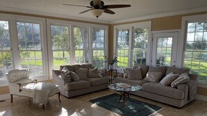 Amplia sala de estar con ventanas grandes, ventilador de techo y una variedad de asientos que incluyen un sofá, sillón y reposapiés.
