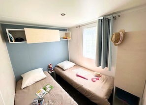A small, well-lit room with two single beds, a shelf above one bed, a window with curtains, and a hat hanging on the wall.