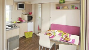 A compact kitchen with a gas stove, microwave, and sink, a dining table with pink cushions, and a small refrigerator. - ZEcamping 4 étoiles 200m plage piscine couverte club enfant mobil-home TV (La Faute-sur-Mer)