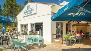 An outdoor seating area with tables and chairs, a white building with a sign reading "LES PLOTS BLEUS," and a blue awning with "MIGUELA" written on it. - ZEcamping 4 étoiles 200m plage piscine couverte club enfant mobil-home TV (La Faute-sur-Mer)