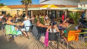 A crowded outdoor dining area with tables and chairs, a palm tree, and a building with a tiled roof. - ZEcamping 4 étoiles club enfant parc aquatique sauna hammam chiens acceptés (Saint-Georges-d'Oléron)