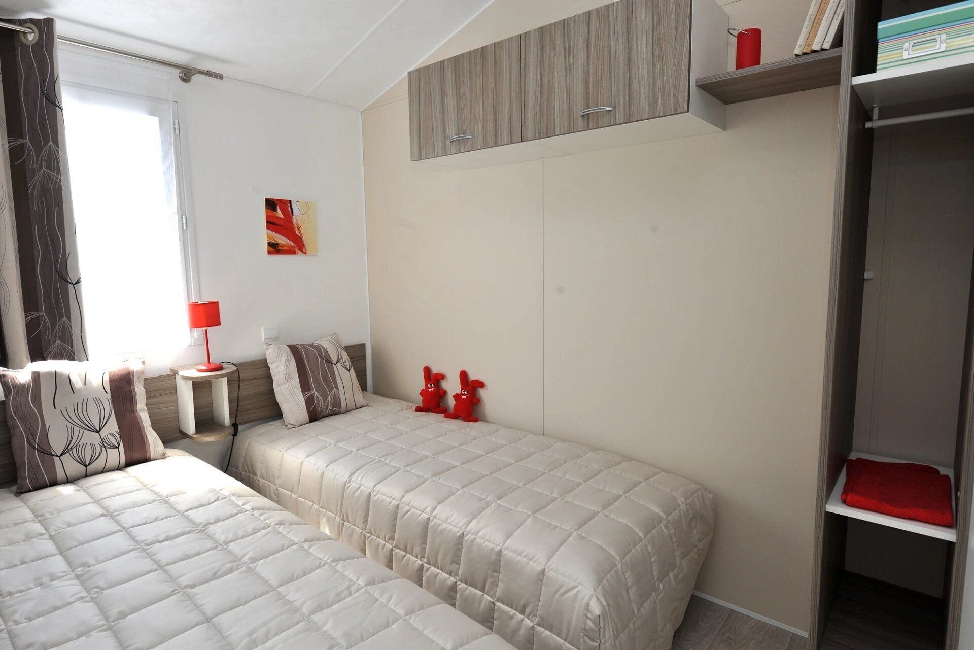 Quarto de hotel com duas camas, um guarda-roupa, uma mesinha com abajur e uma janela com cortinas.