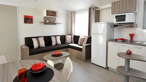 Un espace de vie compact comprenant un canapé, une table à manger, un coin cuisine avec réfrigérateur et micro-ondes, et une petite fenêtre avec des rideaux.