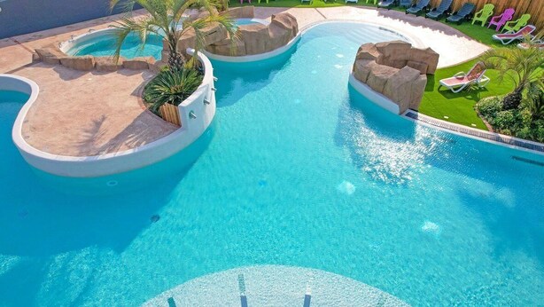 A large, clear swimming pool with multiple sections and a central island, surrounded by lounge chairs and greenery.