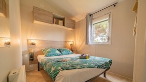 Un dormitorio con cama, mesita de noche, repisa sobre la cama, ventana con cortinas e iluminación en la pared.