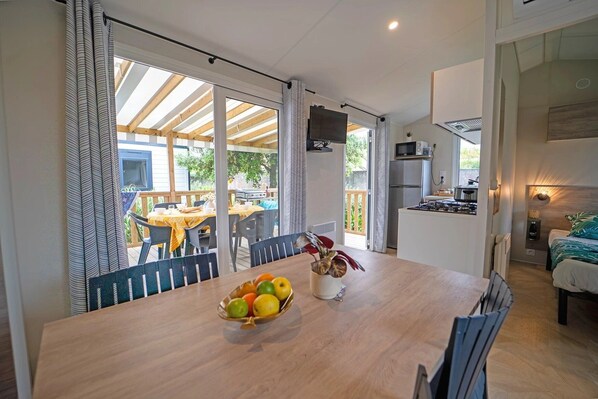 A dining area with a wooden table, chairs, and a fruit bowl. There is a kitchen area with a stove and a microwave. The room has large windows with curtains, and a balcony is visible outside.