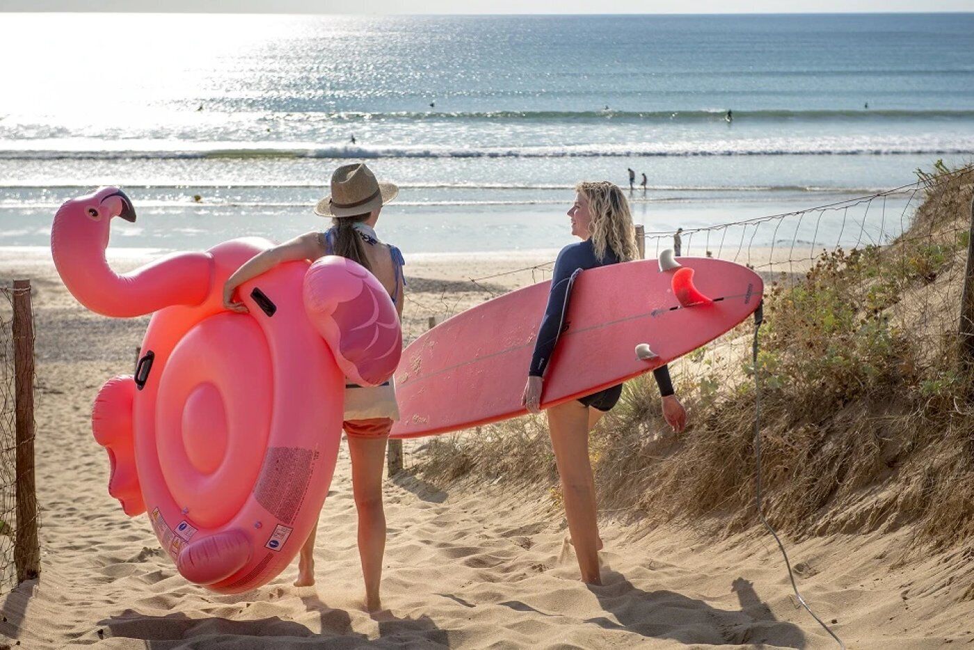 Two people on a beach with a pink flamingo float and a red surfboard.