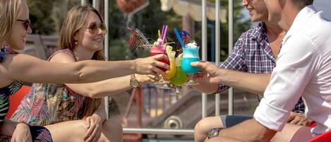 A group of people toasting with colorful drinks at a poolside setting.