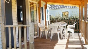 Un espacio de estar al aire libre cubierto con sillas de plástico blanco y una mesa, suelo de madera y vistas a un edificio azul claro con tejado de tejas.