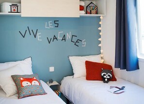 A bedroom with two beds, each with white bedding and a variety of pillows, including a fox and a raccoon design. The wall above the beds has the phrase "LES VIVE VAENCES" written in black letters. There is a shelf with various items and a window with curtains.