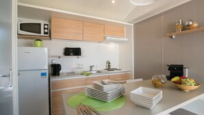 Une cuisine compacte équipée d’un four à micro-ondes, d’un réfrigérateur et d’une petite machine à café. On y trouve des placards en bois, un évier et un panier à fruits sur le plan de travail.