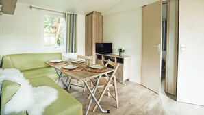 Ein gemütlicher Wohnraum mit einem grünen Sofa, einem Esstisch gedeckt mit Tellern und Gläsern, einem Fernseher auf einem Holzgestell und einer Tür, die in einen anderen Raum führt.