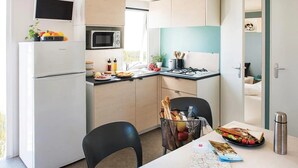 A compact kitchen with a microwave, stove, and refrigerator. There is a dining table with chairs, a basket of bread, and a water bottle.