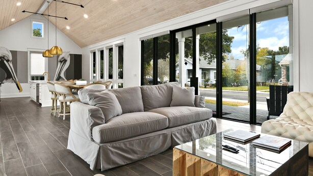 A modern living room with a grey sofa, a glass coffee table, and a white armchair. There is a dining area with a table and chairs, and large windows offering a view of the outside. - The Luxe on Main is an Ultra-Modern, Trendy New Construction in Downtown BSL. (Bay St. Louis)