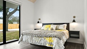 A bedroom with a large bed, two bedside tables with lamps, a sliding glass door, and a view of the outdoors. - The Luxe on Main is an Ultra-Modern, Trendy New Construction in Downtown BSL. (Bay St. Louis)