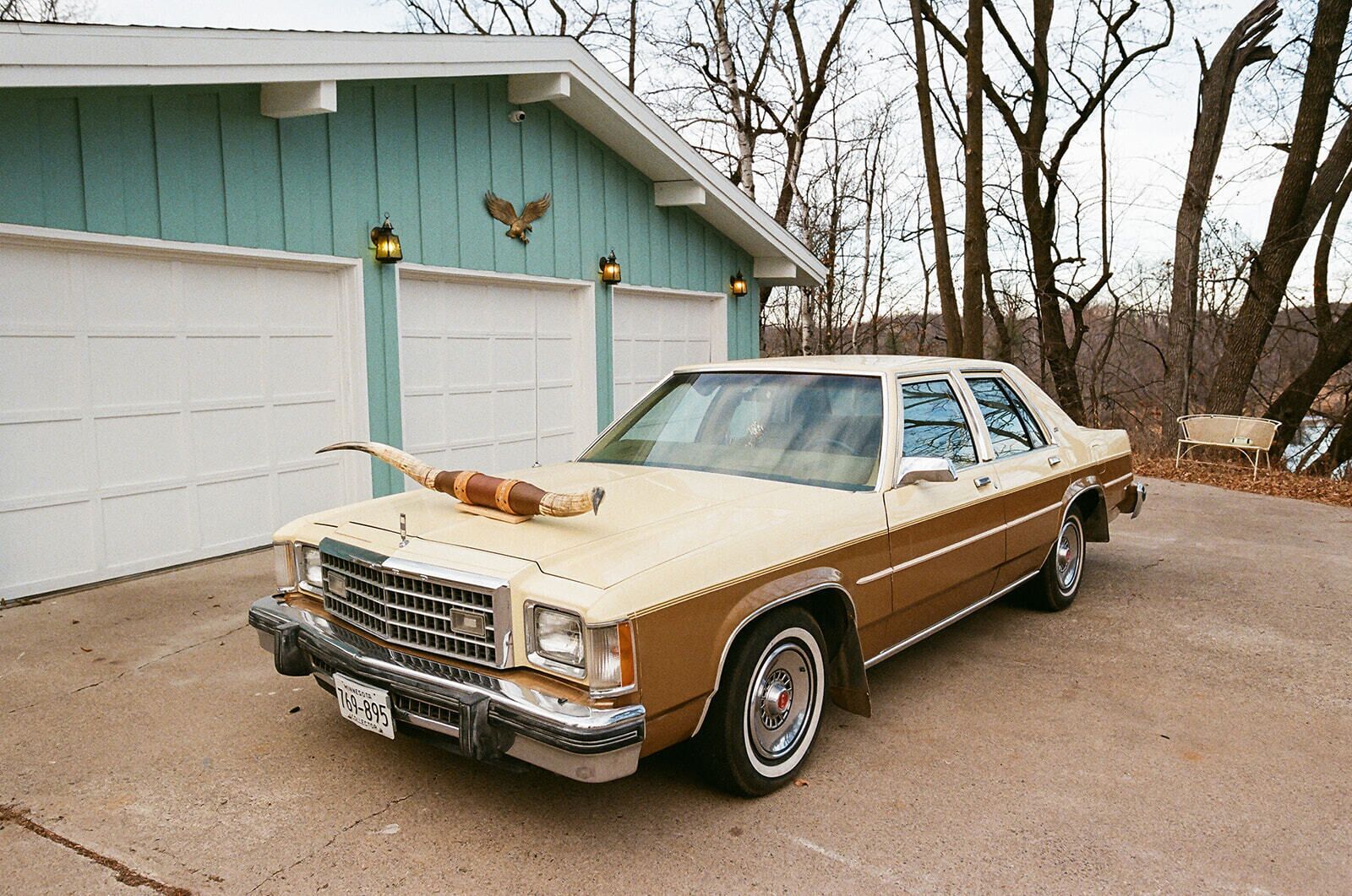 Une voiture ancienne, de couleur beige et marron, est garée devant un garage dont la façade est turquoise et la porte blanche.