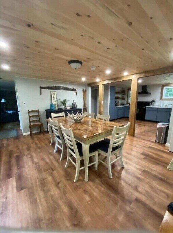 A dining area with a wooden table and chairs, a kitchen in the background, and a ceiling with wooden beams.