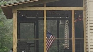 Una cabaña de madera con balcón, una bandera estadounidense y un fondo boscoso.