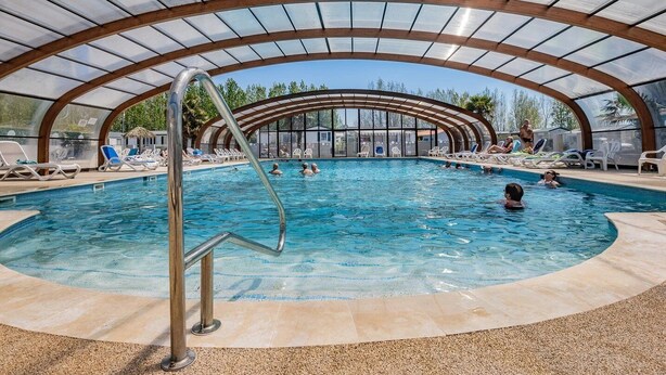 An indoor swimming pool with a glass ceiling, surrounded by lounge chairs and a poolside area.