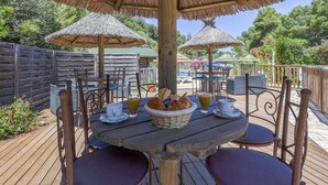 Un área de comedor al aire libre con mesas y sillas de madera, sombrillas de paja y una piscina al fondo.
