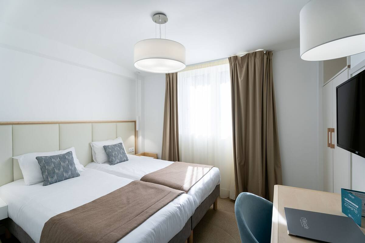 Quarto de hotel com duas camas, uma escrivaninha, televisão e uma janela grande com cortinas.