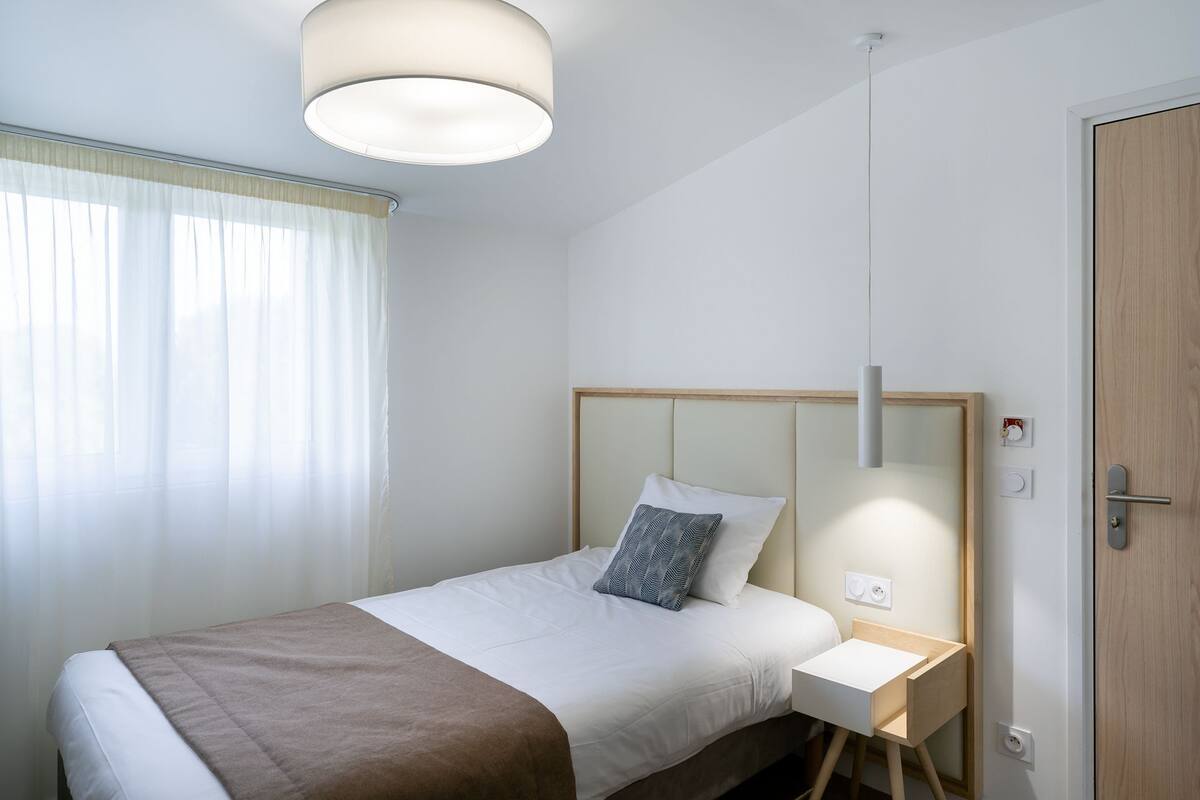 Uma cama bem arrumada com um cobertor marrom, uma mesinha de cabeceira com abajur e uma porta de madeira.