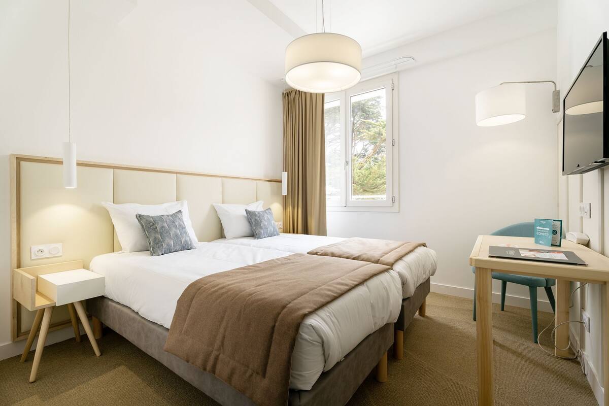 Quarto de hotel com cama de casal, escrivaninha com cadeira, televisão e janela com cortinas.