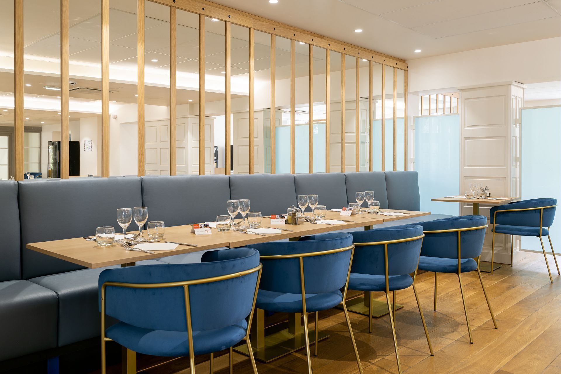 A restaurant with a long table set for dining, blue upholstered seating, and a wooden partition with glass panels.