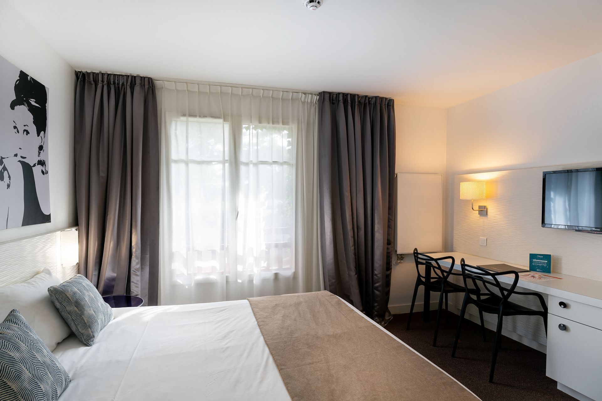 Hotelkamer met een bed, een bureau en een stoel. Er is een raam met gordijnen, een tv aan de muur en een schilderij.