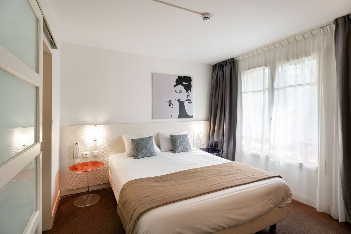 Quarto de hotel com uma cama grande, uma mesa de cabeceira, luminária na parede, janela com cortinas e uma câmera de segurança.