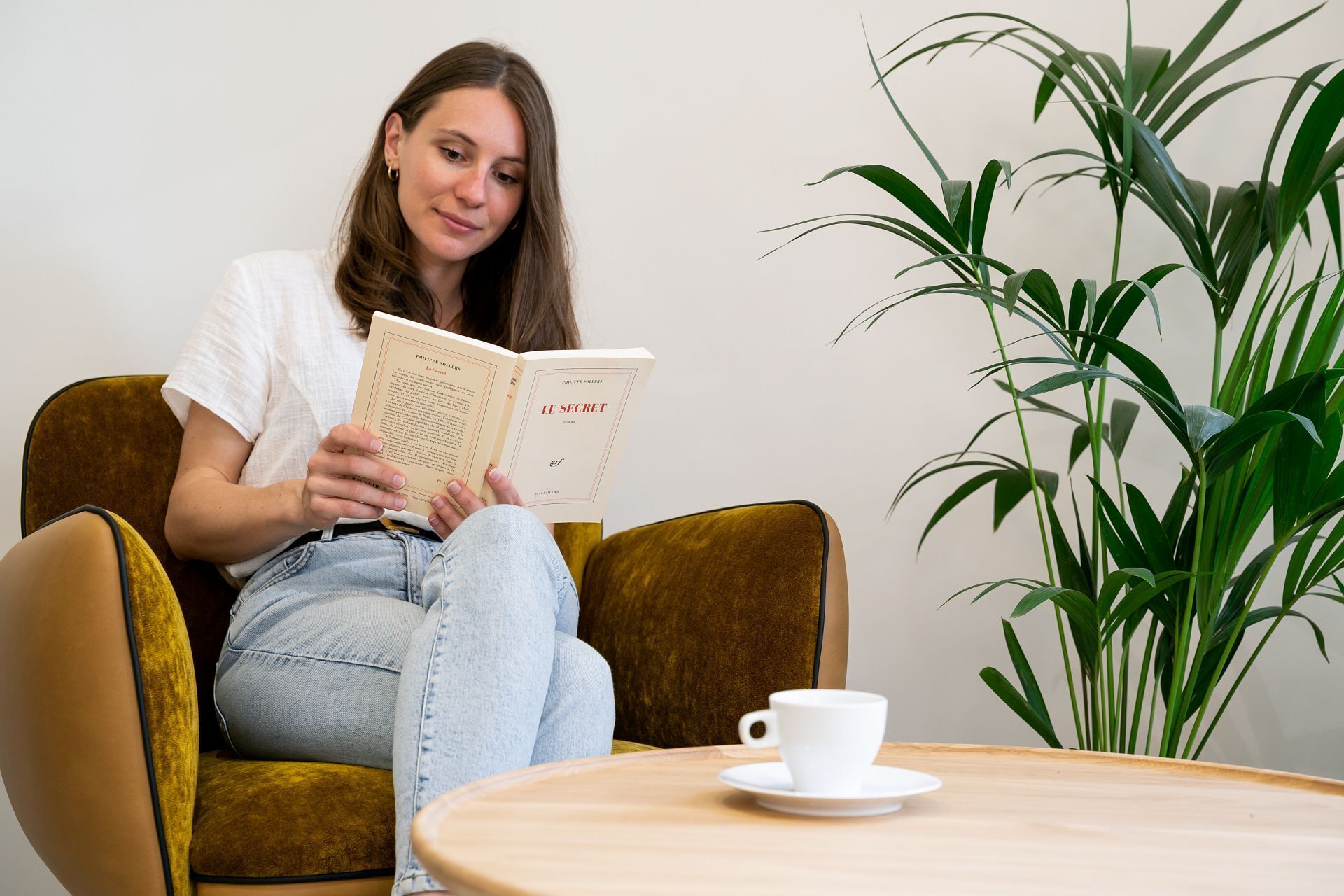 Eine Person sitzt in einem senffarbenen Sessel und liest ein Buch. Auf einem Holztisch daneben steht eine weiße Tasse.