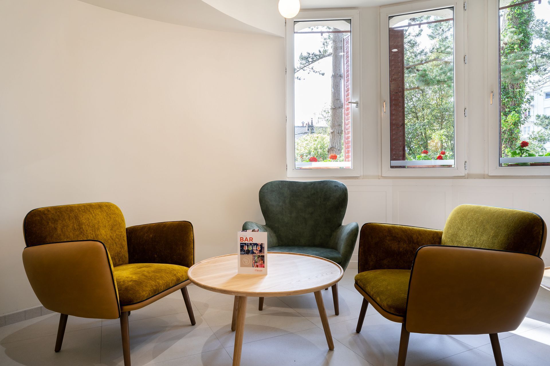 A room with three chairs and a round table, a window with a view of trees and flowers, and a door with a brick wall.