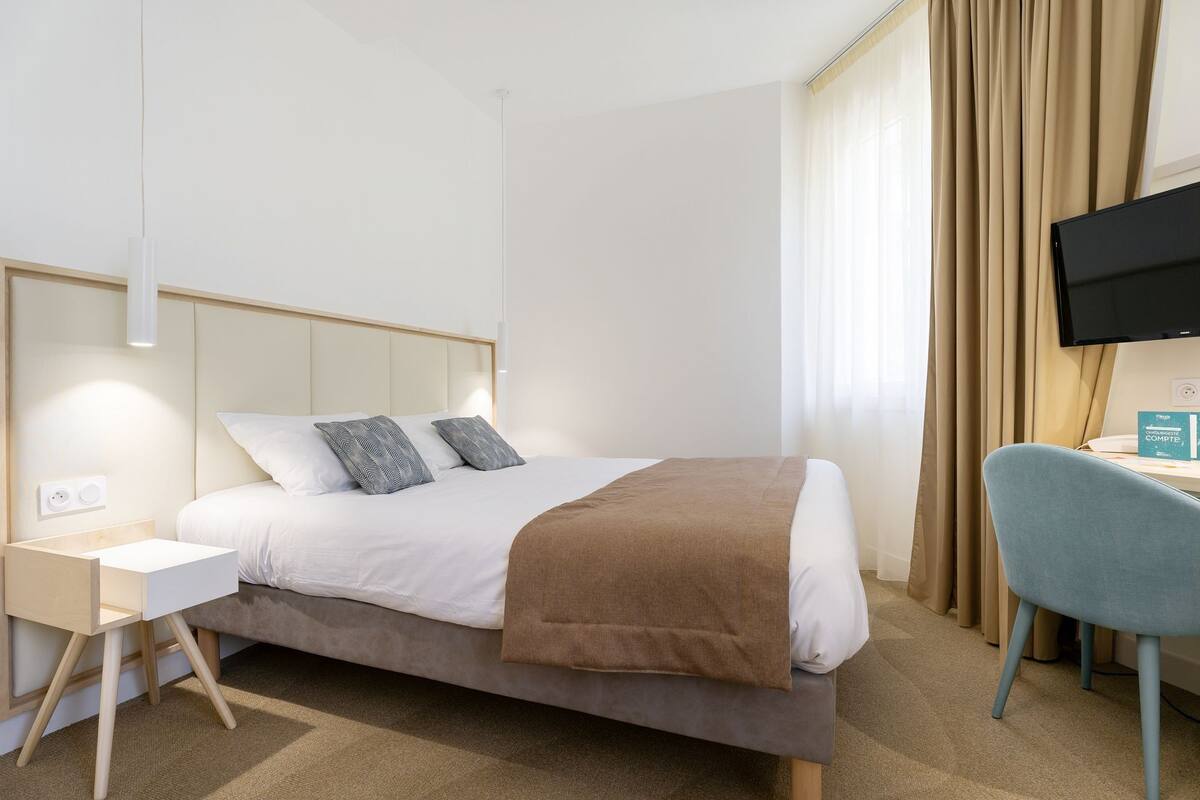 Quarto de hotel com cama, escrivaninha, cadeira, televisão e janela com cortinas.