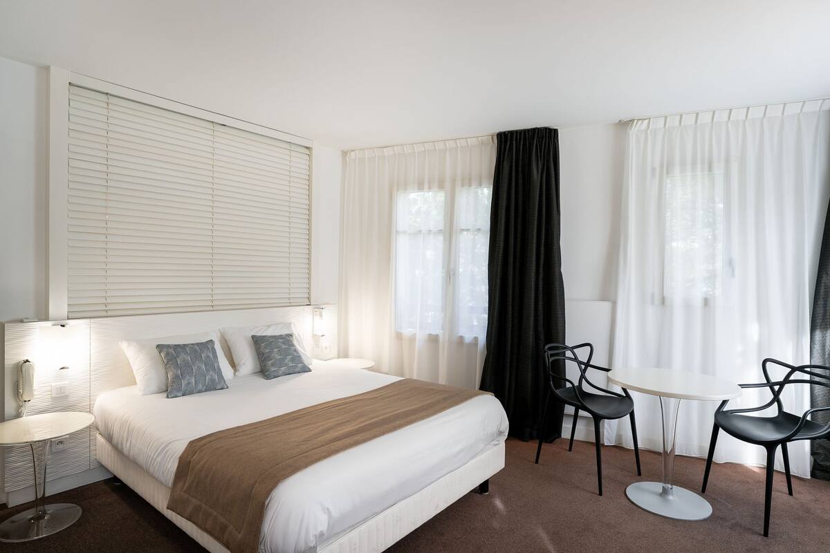 Quarto de hotel com uma cama grande, mesa de cabeceira, cadeira, mesa redonda pequena e uma janela com cortinas.