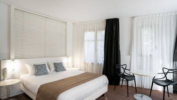 A hotel room with a large bed, a bedside table, a chair, a small round table, and a window with curtains.