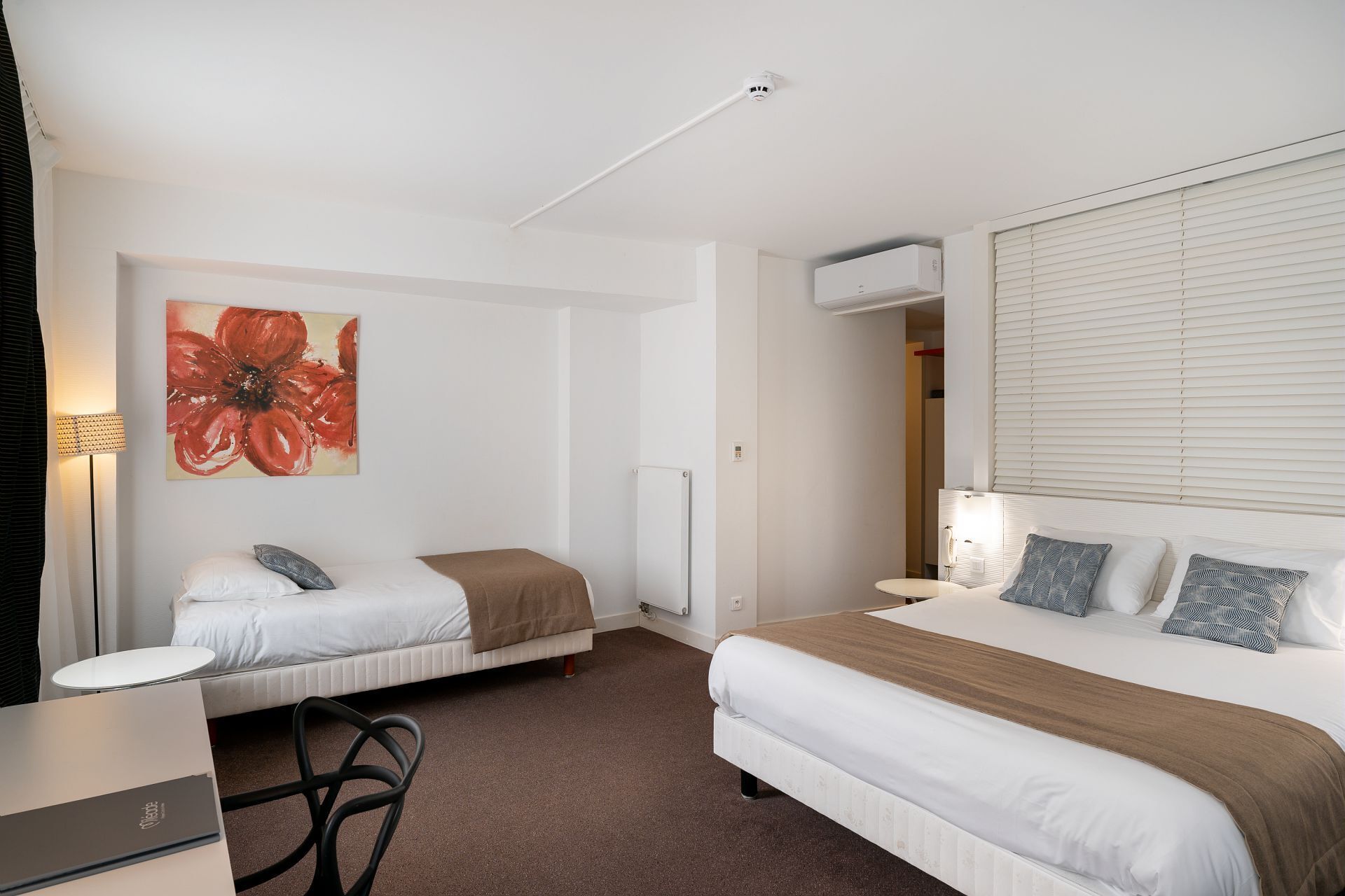 Habitación de hotel con dos camas, un escritorio, una silla, un radiador y un ventanal grande con persianas.