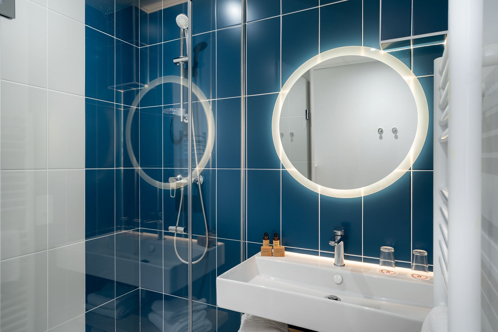 Un baño moderno con ducha envuelta en cristal, un lavabo blanco con espejo y paredes de azulejos azules.