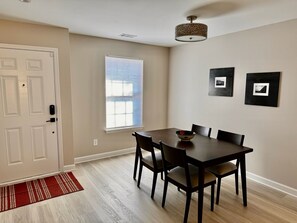 Un coin repas avec une table et des chaises, une fenêtre avec des stores, une porte avec une serrure et un tapis rayé rouge et blanc.
